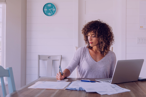 Woman reviewing paperwork at a kitchen table with a laptop beside her.